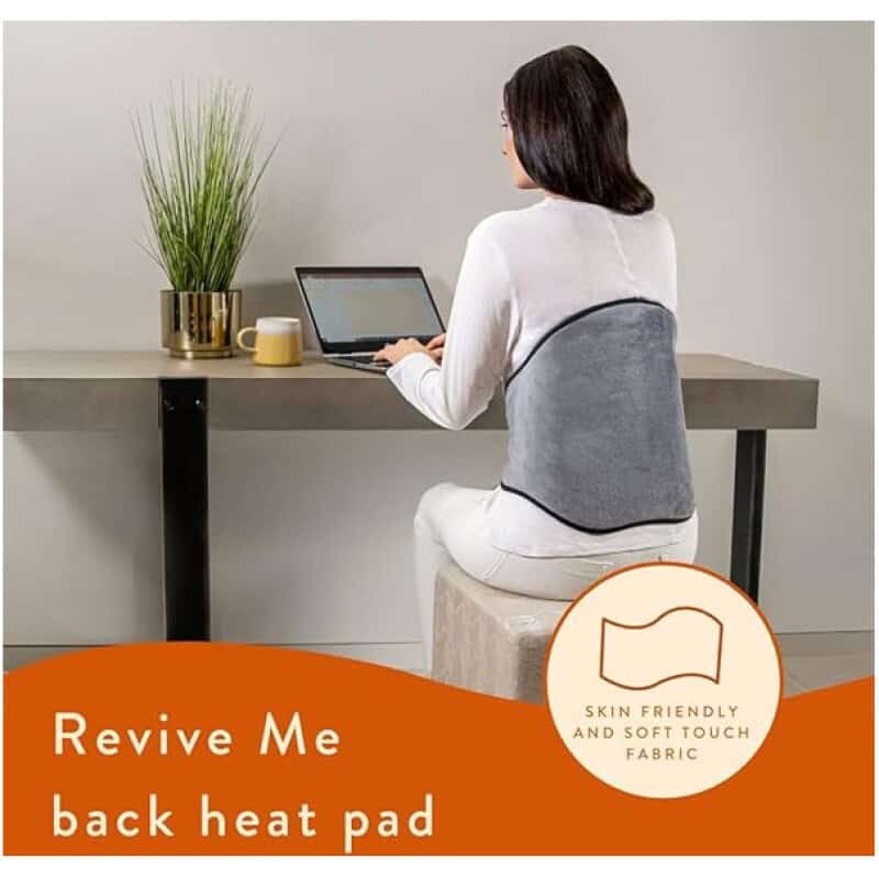 Back view of woman working on laptop at desk with heat pad on her back, promoting comfortable ergonomic office setup.